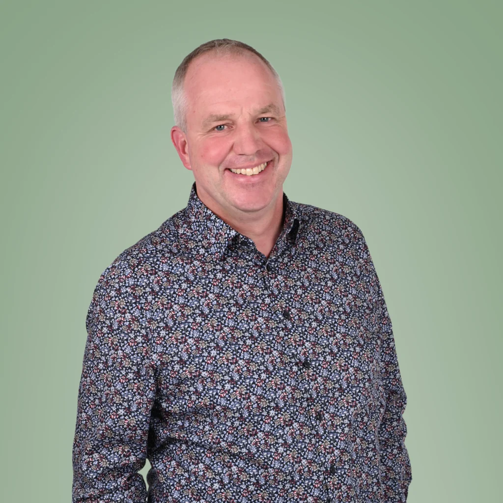 Eine lächelnde Person in einem floralen Hemd vor einem hellgrünen Hintergrund.