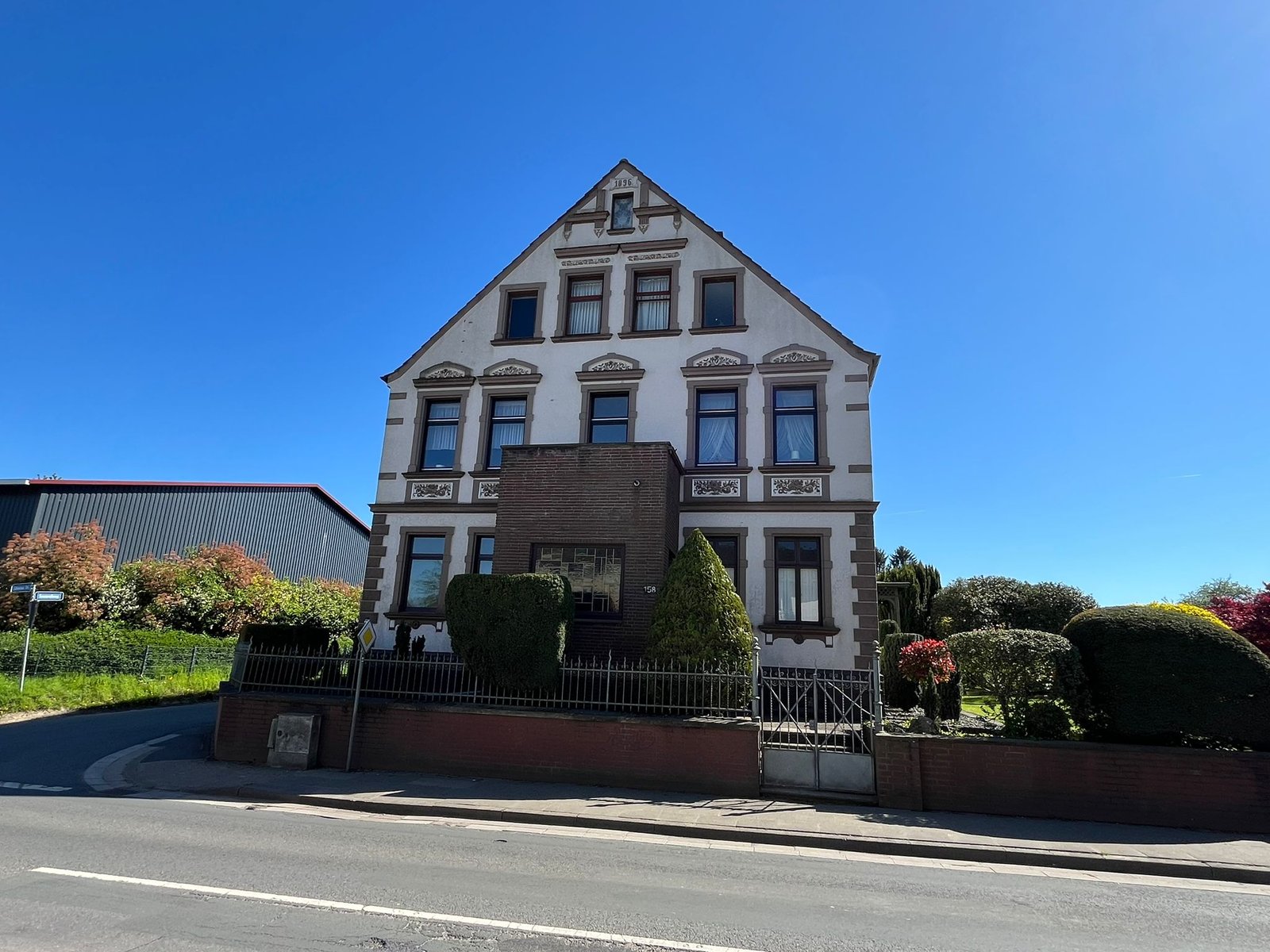Traditionelles weißes Haus mit Garten, umgeben von Zaun, unter klarem Himmel in ruhiger Wohngegend.